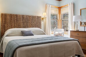 A neatly made bed with a patterned headboard, a bedside table with a lamp, and a window with curtains. - Beachfront, Just steps and you're on the beach! (Saco)