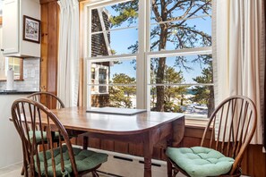 A dining area with a wooden table and chairs, large windows with curtains, and a view of trees and a building outside.