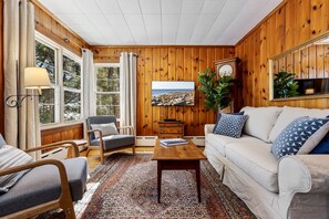 A cozy living room with wooden walls, a large window, a sofa, a chair, a coffee table, and a clock.