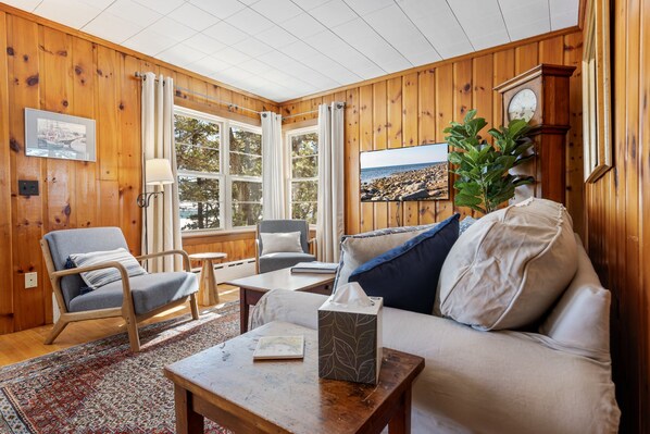 A cozy room with wooden walls, a sofa, a chair, a table, and a window with curtains.