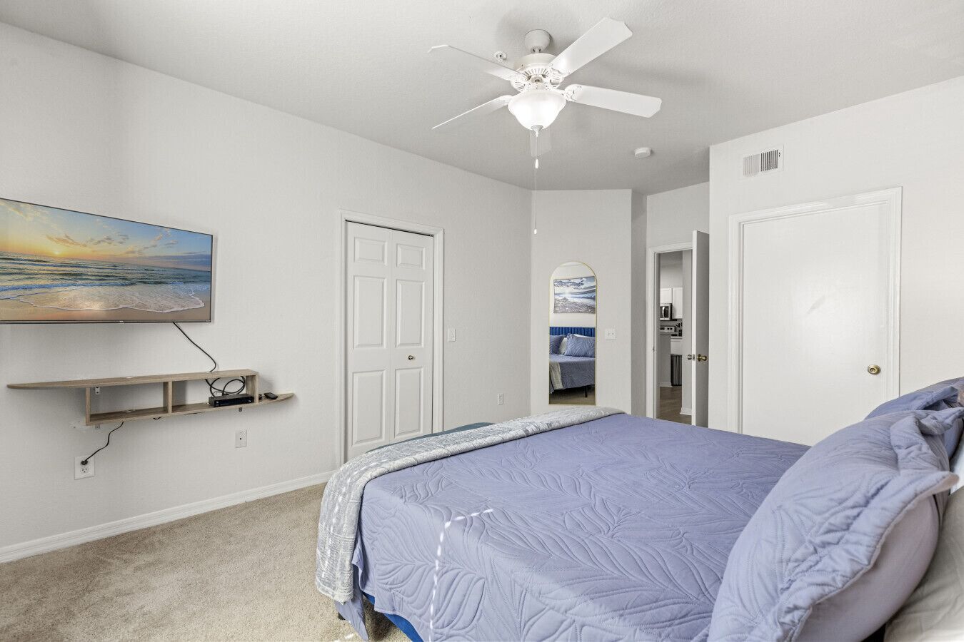 A bedroom with a bed, a ceiling fan, a TV mounted on the wall, and a door leading to another room.