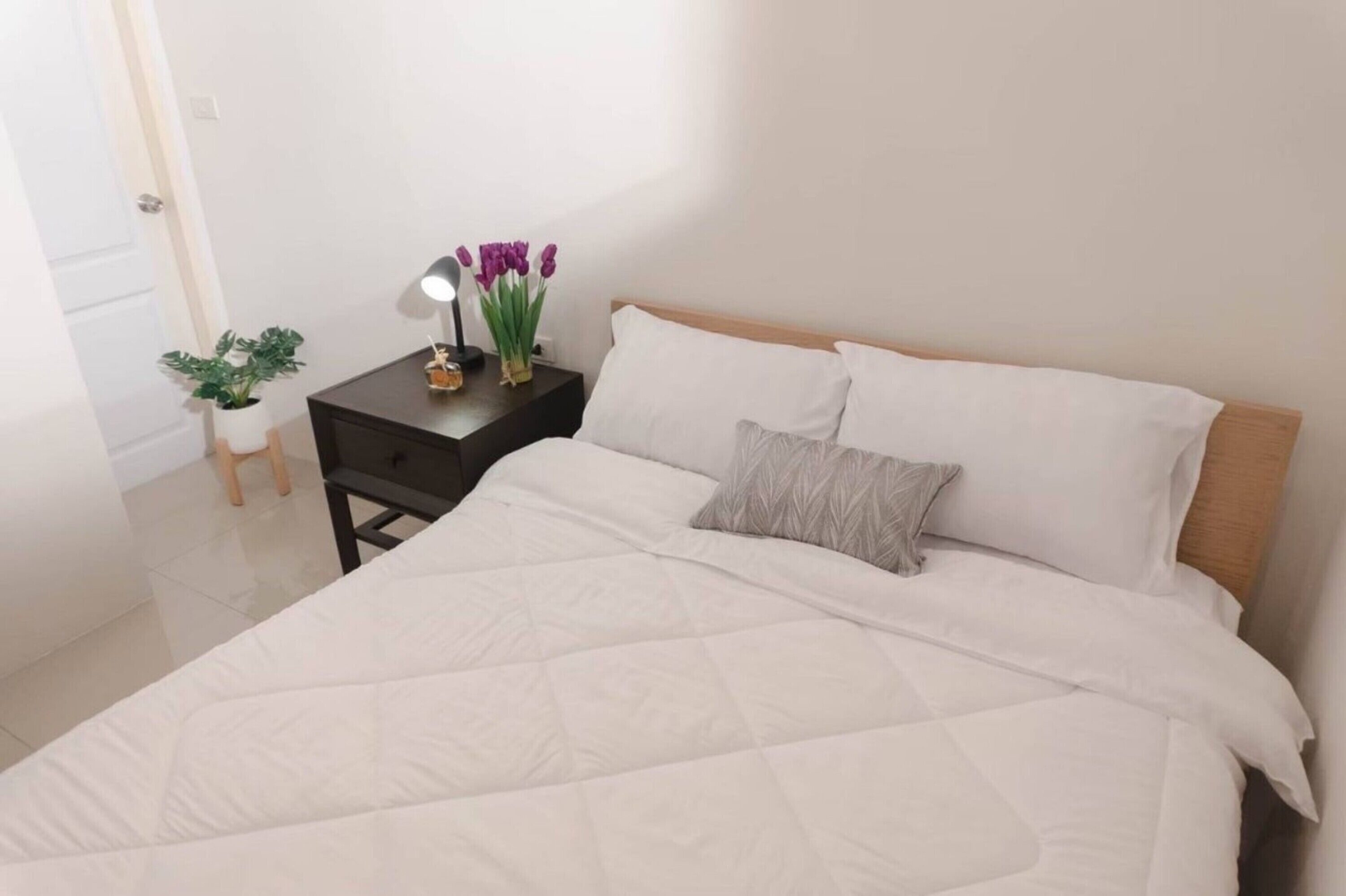 Una cama bien tendida con ropa de cama blanca, una mesita de noche con una lámpara y una planta en maceta.