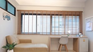 Una habitación minimalista con sofá, escritorio, silla, refrigerador pequeño y ventana con persianas.