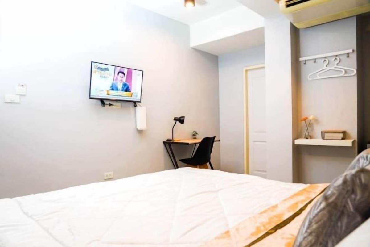 A bedroom with a bed, a desk with a chair, a TV mounted on the wall, and a door.