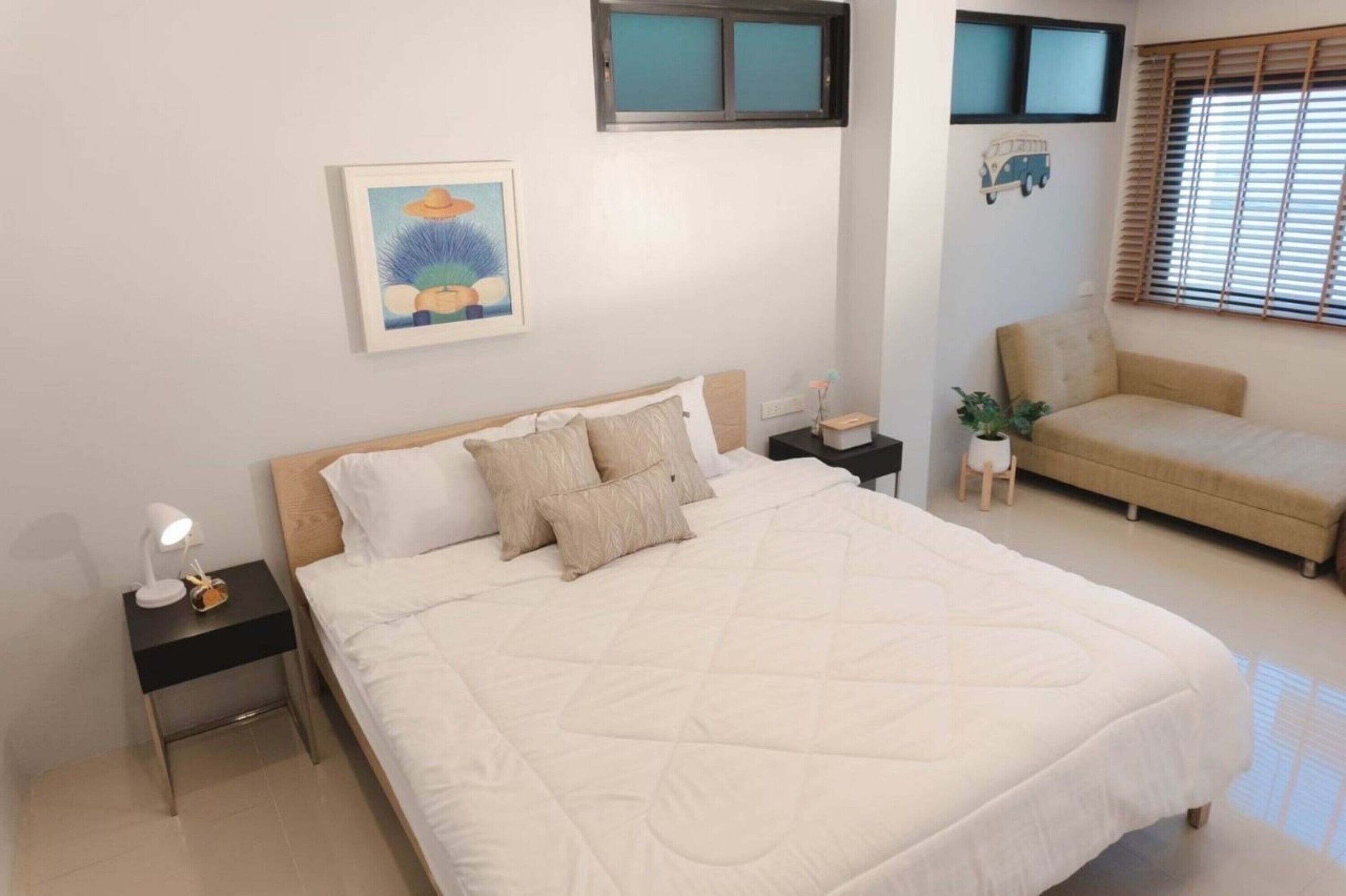 A bedroom with a large bed, a bedside table with a lamp, a small table with a plant, and a window with blinds.