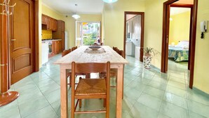 Ruime eethoek met een lange tafel en stoelen, een keuken op de achtergrond en uitzicht op het buitenleven door een open deur.