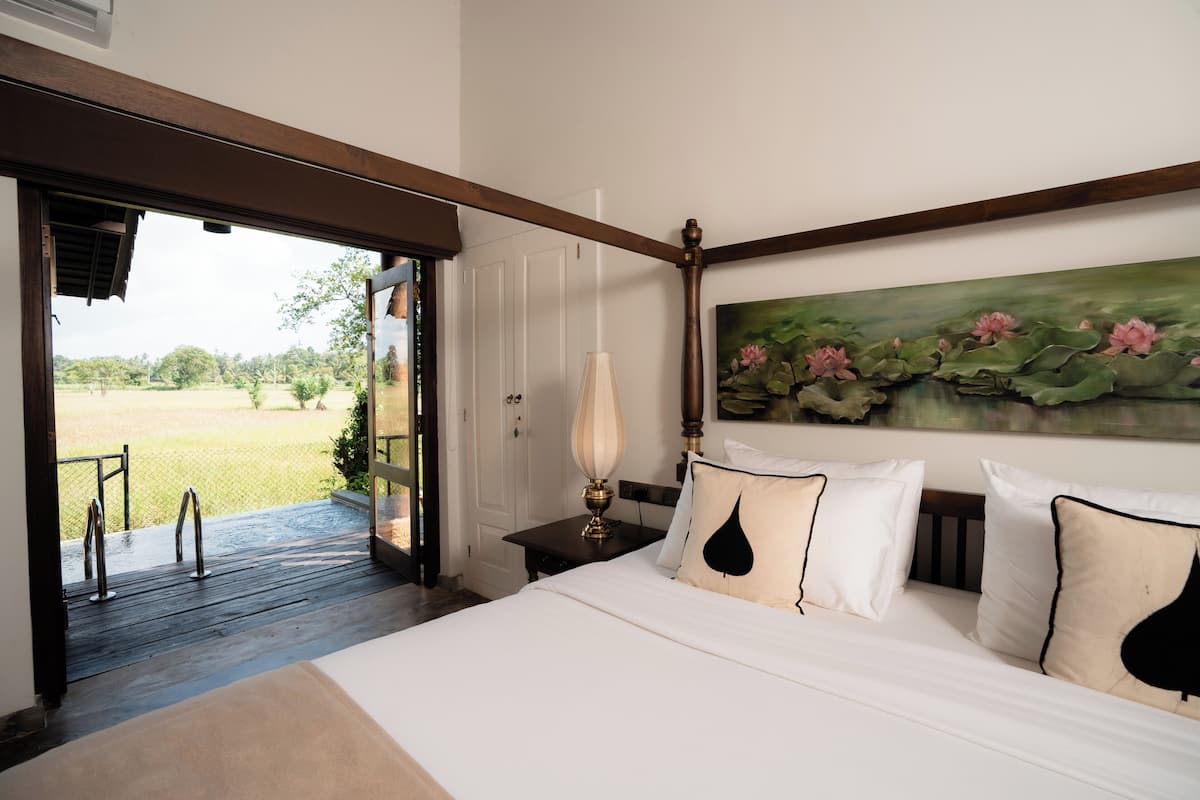 Un dormitorio con cama, almohadas, una mesita de noche, una lámpara y un cuadro de flores de loto.