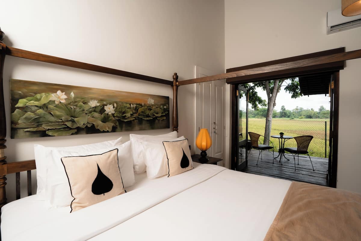 Un dormitorio con una cama grande, un cuadro de nenúfares, cabecero de madera, mesita de noche con lámpara y vistas a una terraza y jardín a través de una puerta de cristal.
