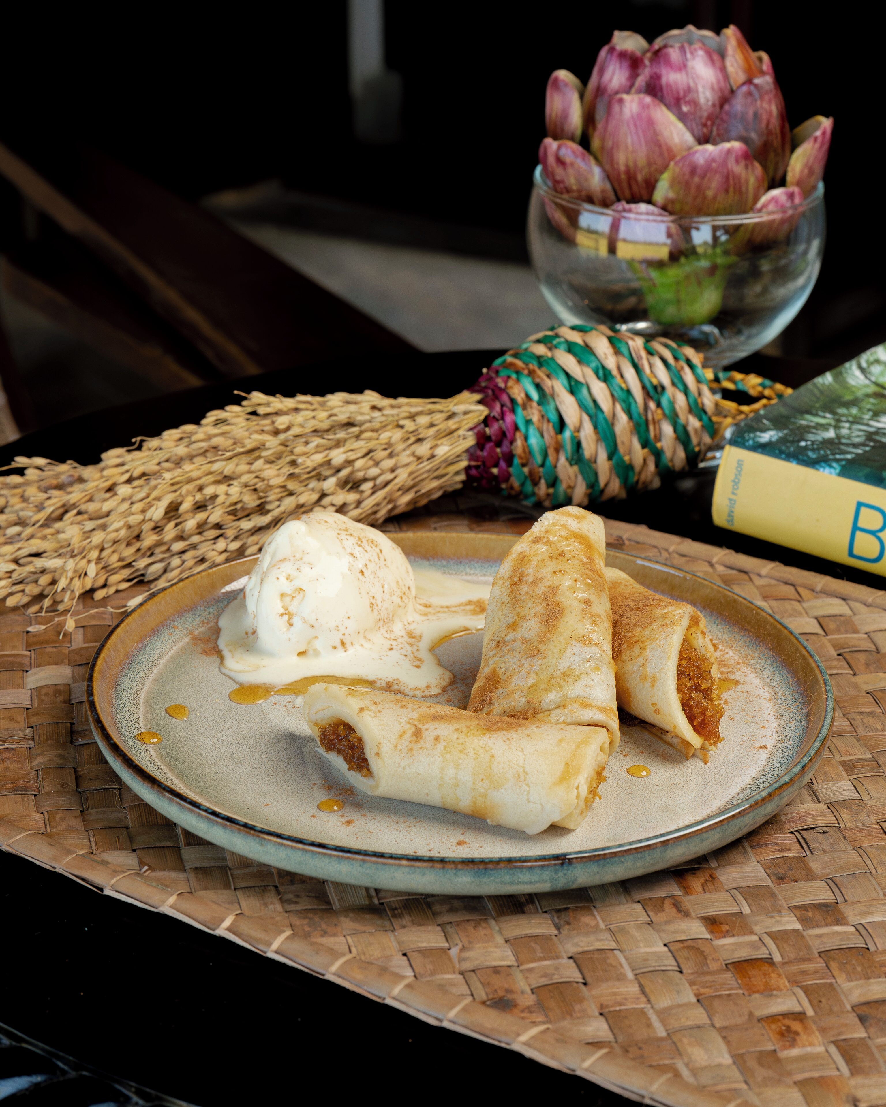A plate with rolled pastries and a bowl of artichokes on a woven mat.