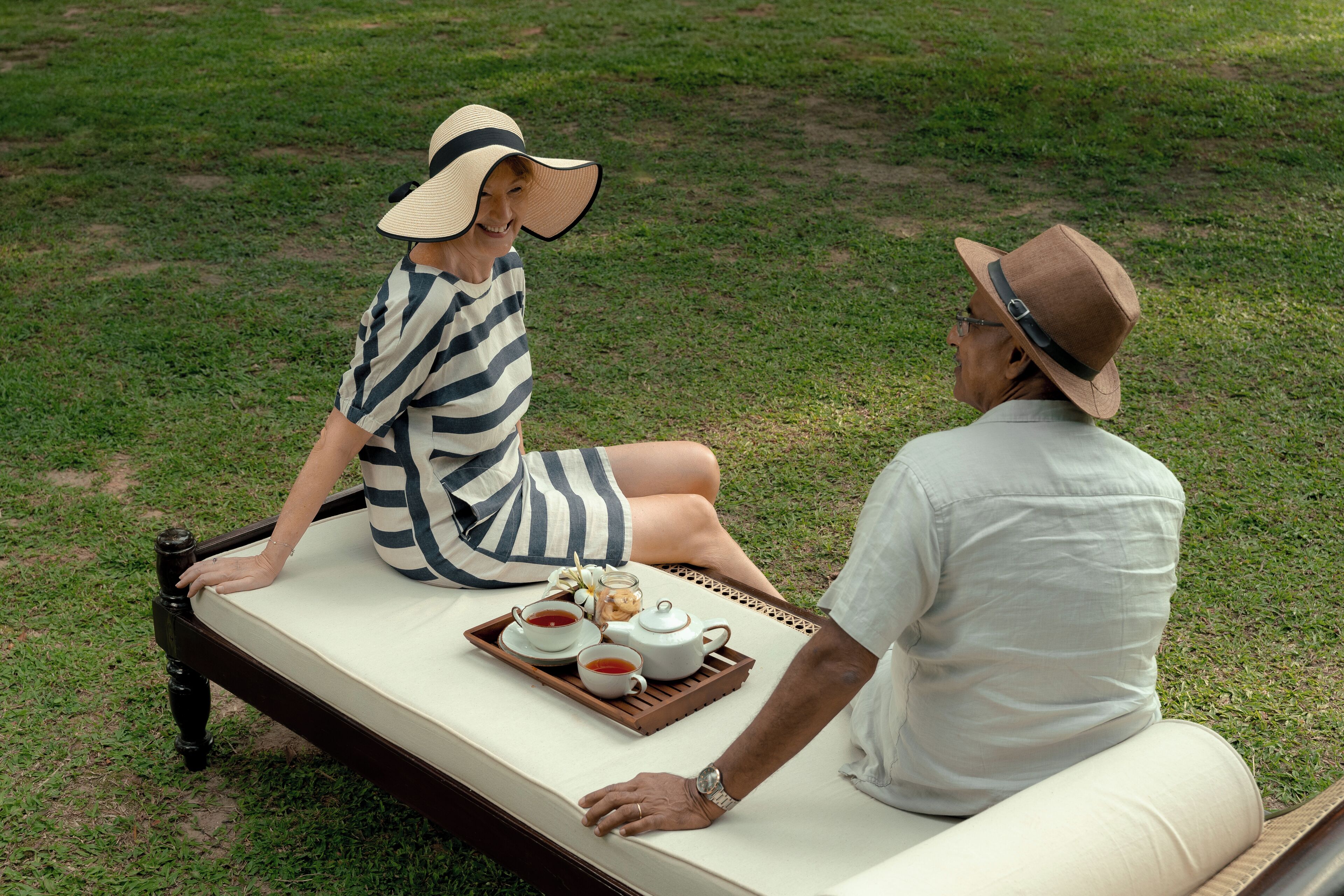 A couple sitting on a bed with a tray of tea and a teapot.