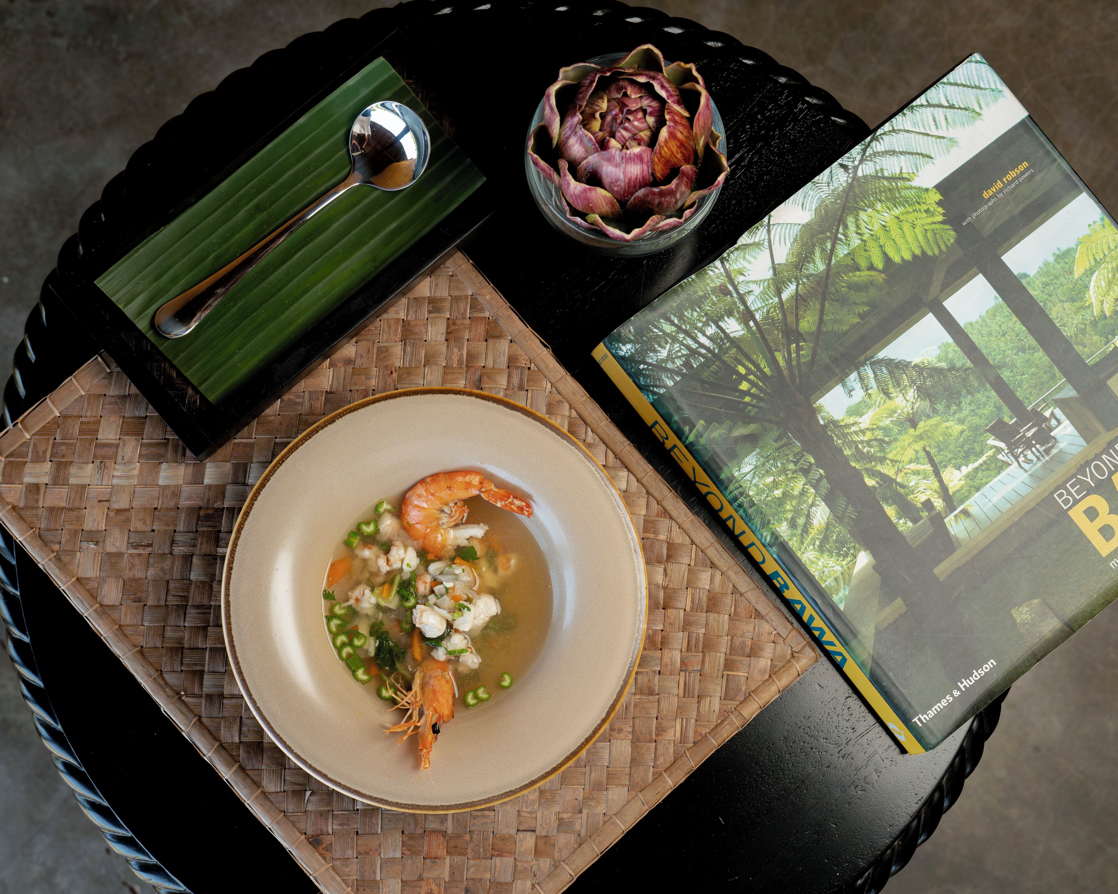 A table with a woven placemat, a bowl of soup with shrimp, a spoon, a decorative bowl, and a book titled "BEYOND B".
