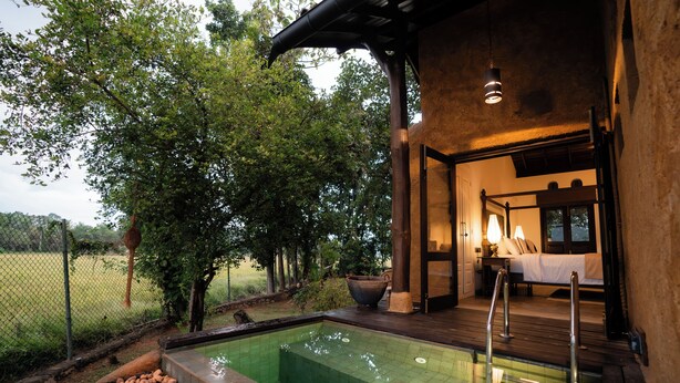 A pool area with a wooden deck, a bed with a white bedspread, and a large window overlooking a garden.