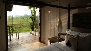 A bedroom with a bed, bedside table, lamp, and a view of a green landscape through an open door.