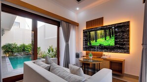 A modern living room with a large flat-screen TV, a sofa, a small table, and a pool visible through the sliding glass doors. - Villa Boswelia Seminyak (Seminyak)