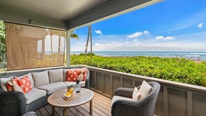 A covered outdoor seating area with a view of the ocean, a sofa, two chairs, and a small round table with a fruit bowl.