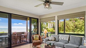 A spacious living area with a large sliding glass door leading to a balcony with a view of the ocean. Inside, there is a gray sofa, a wooden coffee table, and a wicker side table with a lamp.