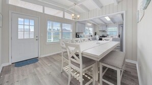 Una cocina amplia con armarios blancos, un comedor con mesa y sillas blancas, y ventanas grandes que dejan entrar luz natural.