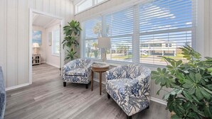 Una habitación con dos sillones estampados, una mesita redonda pequeña y amplias ventanas con persianas.