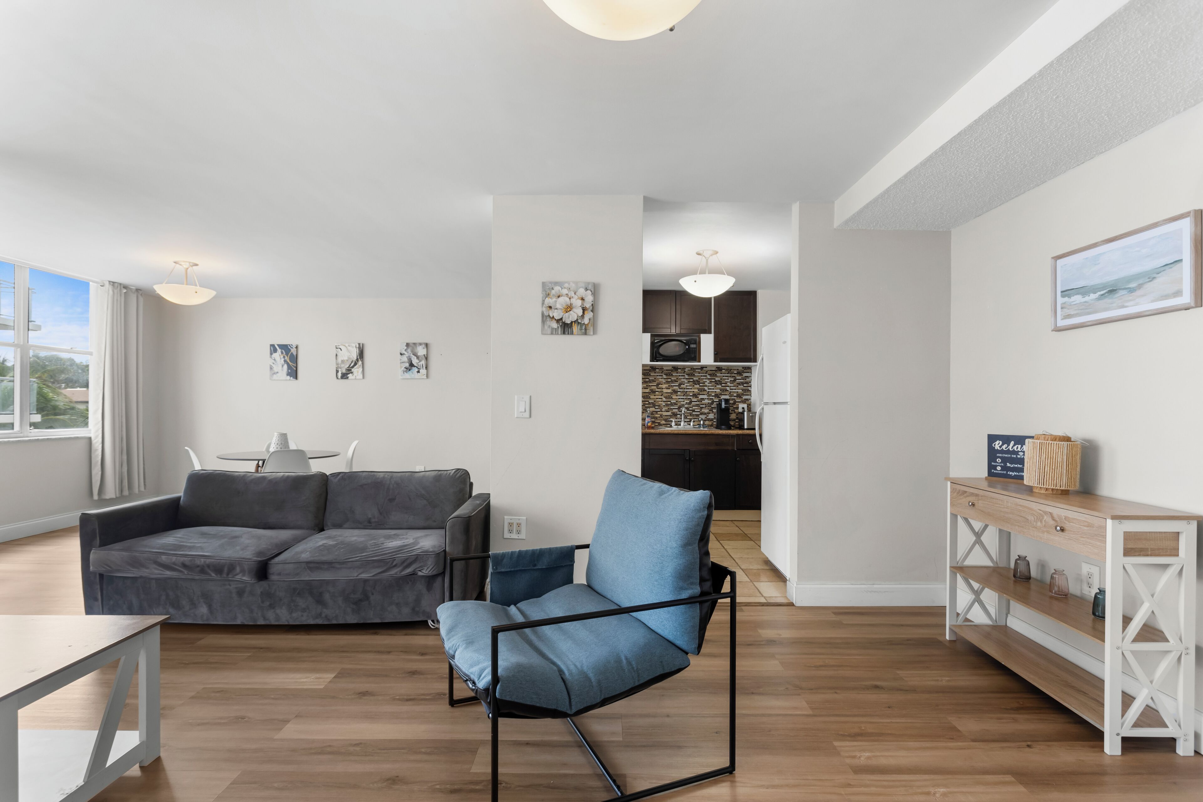 A modern living room with a grey sofa, a blue armchair, a white console table, and a kitchen in the background.
