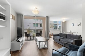 A modern living room with a grey sofa, a white coffee table, and a TV on a white stand. There is a balcony with a table and chairs. - Bay Harbor Sunset Apt 3B (Bay Harbor Islands)