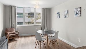 A modern room with a round table, four chairs, a brown armchair, and a window with curtains. - Bay Harbor Sunset Apt 3B (Bay Harbor Islands)