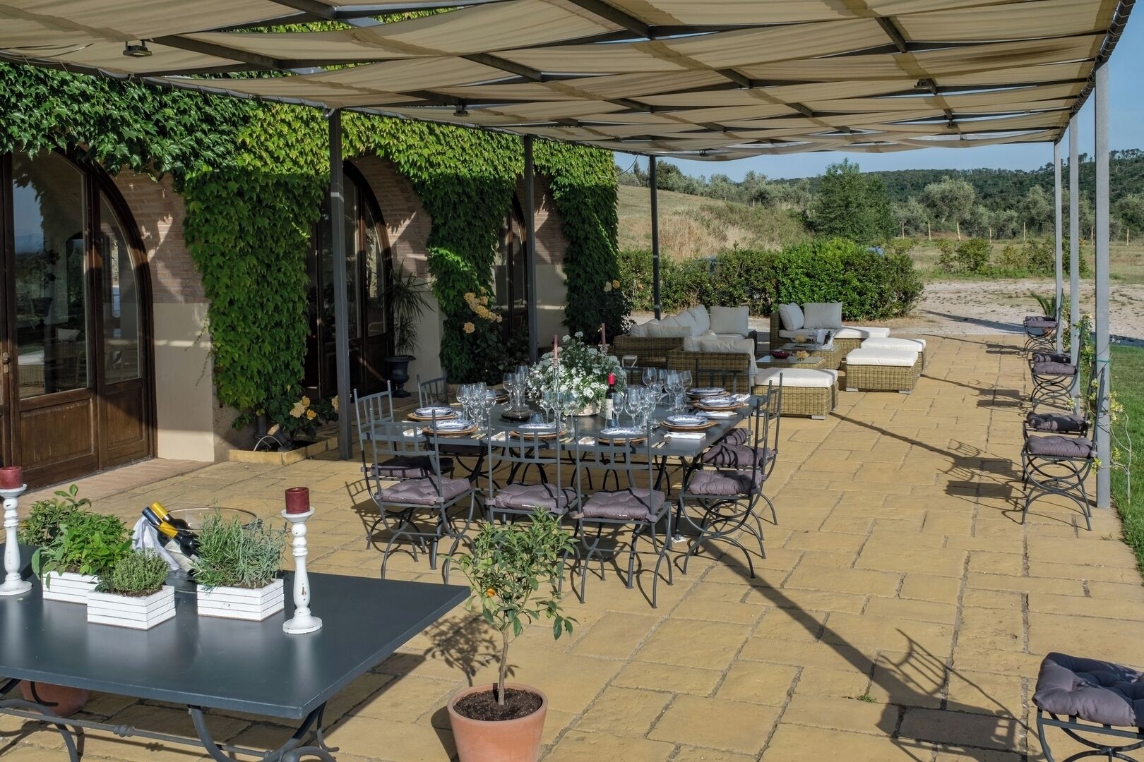 An outdoor patio area with a covered dining table set for six, surrounded by wicker furniture and potted plants.