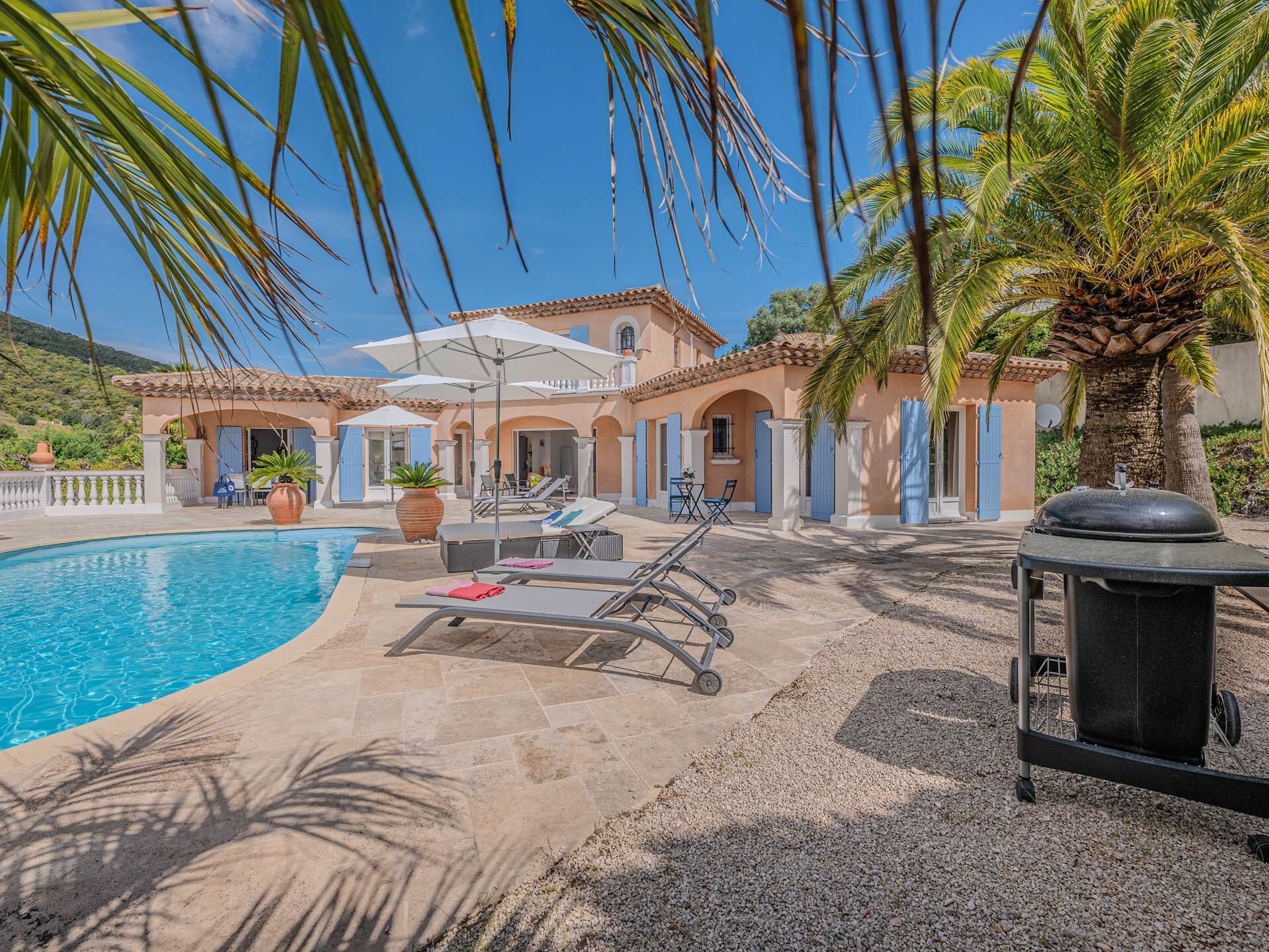 A poolside area with lounge chairs, a barbecue grill, and a building with a tiled roof and arches.