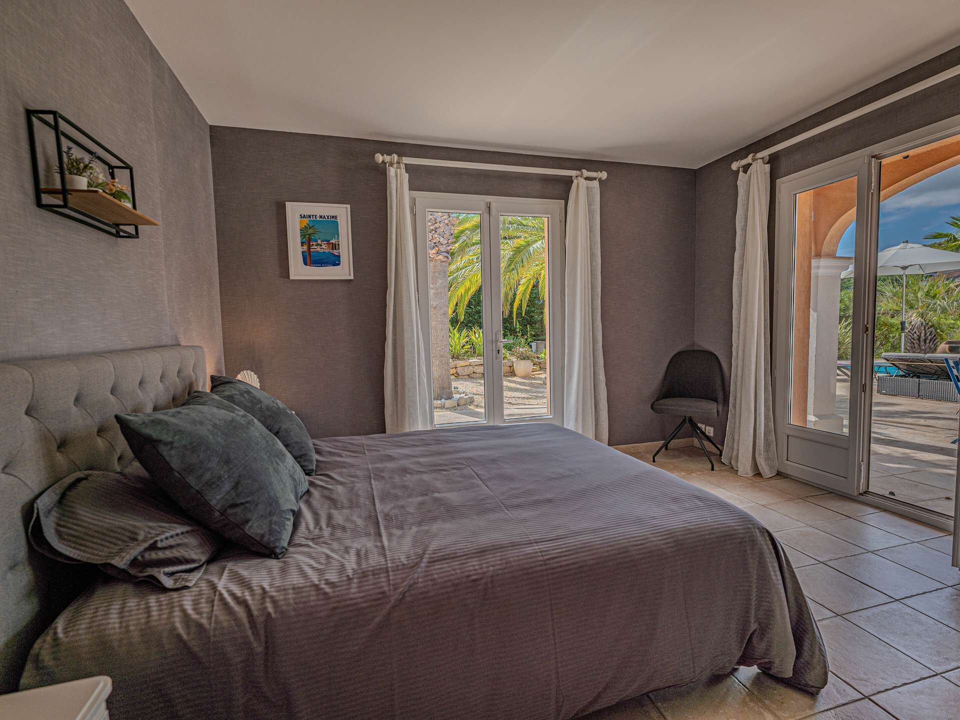 A bedroom with a bed, a chair, a picture on the wall, and a view of the outdoors.