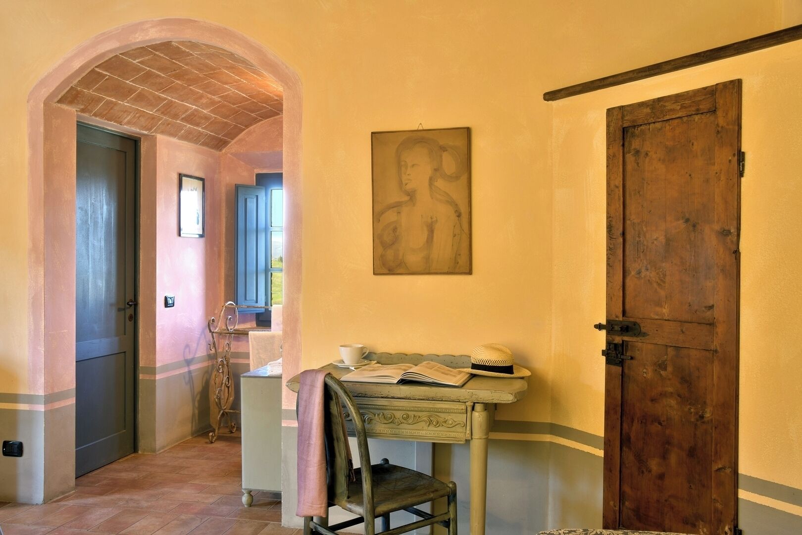 A hallway with a rustic door, a table with a book and a hat, and a painting on the wall.