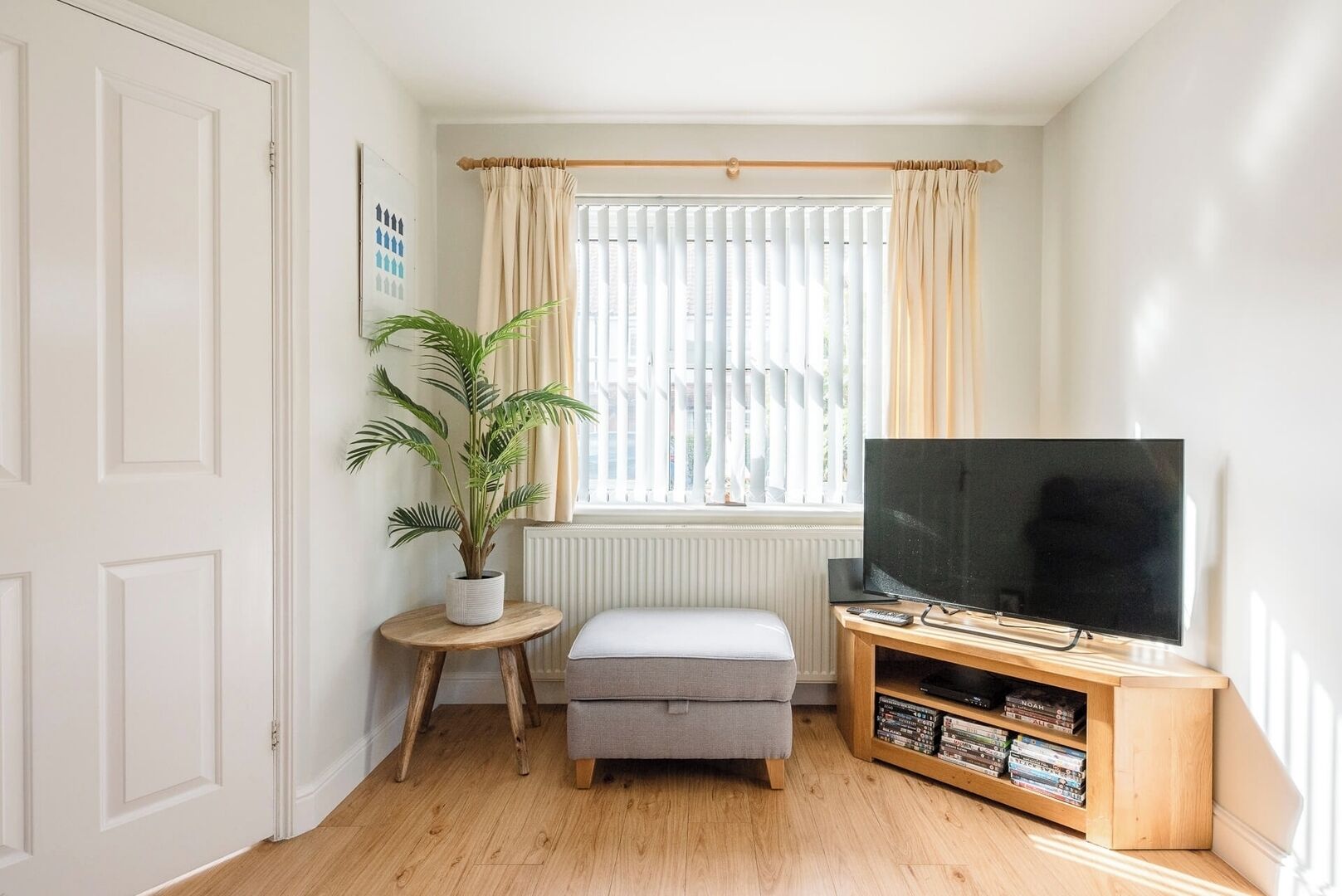 A cozy living room with a television, a small side table with a plant, a cushioned bench, and a wooden entertainment unit with books.