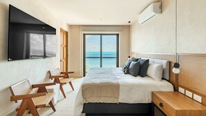 A modern bedroom with a large bed, a TV mounted on the wall, and a view of the ocean through a large window.