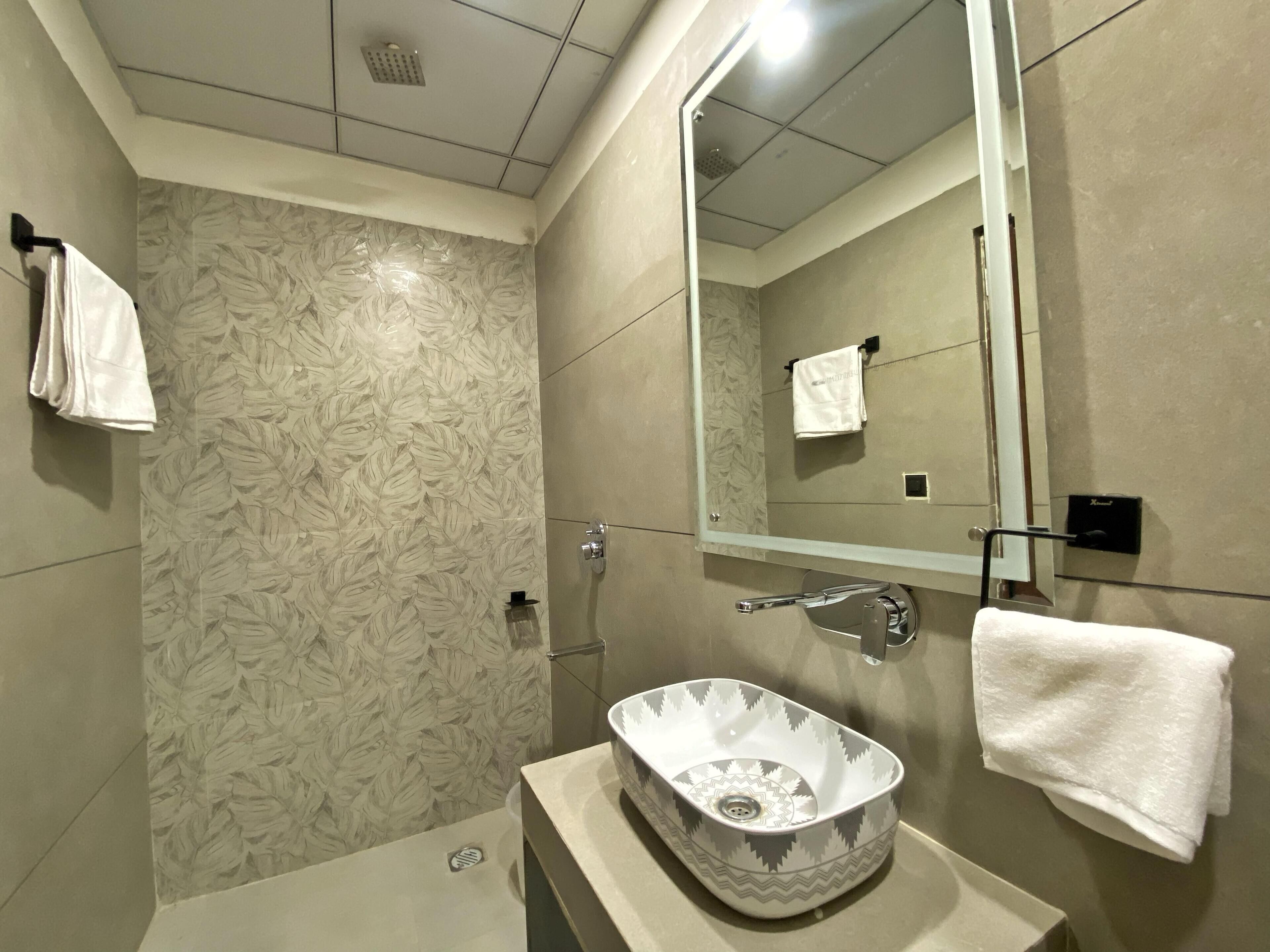 Un baño moderno con ducha, lavamanos con un lavabo decorativo, un espejo grande y un perchero para toallas.