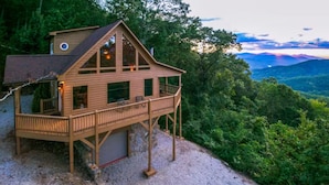 A wooden cabin with a balcony, set in a forested area with a view of mountains.