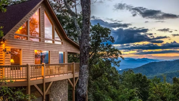 A cabin with a stone foundation, large windows, and a balcony overlooking a mountainous landscape.