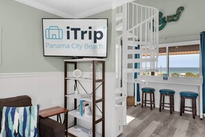 A room with a white wall-mounted sign reading 'iTrip Panama City Beach,' a white shelving unit with decorative items, a wooden bench, and a view of the ocean through a window with a white railing.