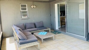 A sunlit balcony with a grey sofa, a wooden table, and a sliding glass door leading to a kitchen area. - Close to Best Gulf Beaches, Downtown Venice & Wellen Park - Long Term Available (Venice)