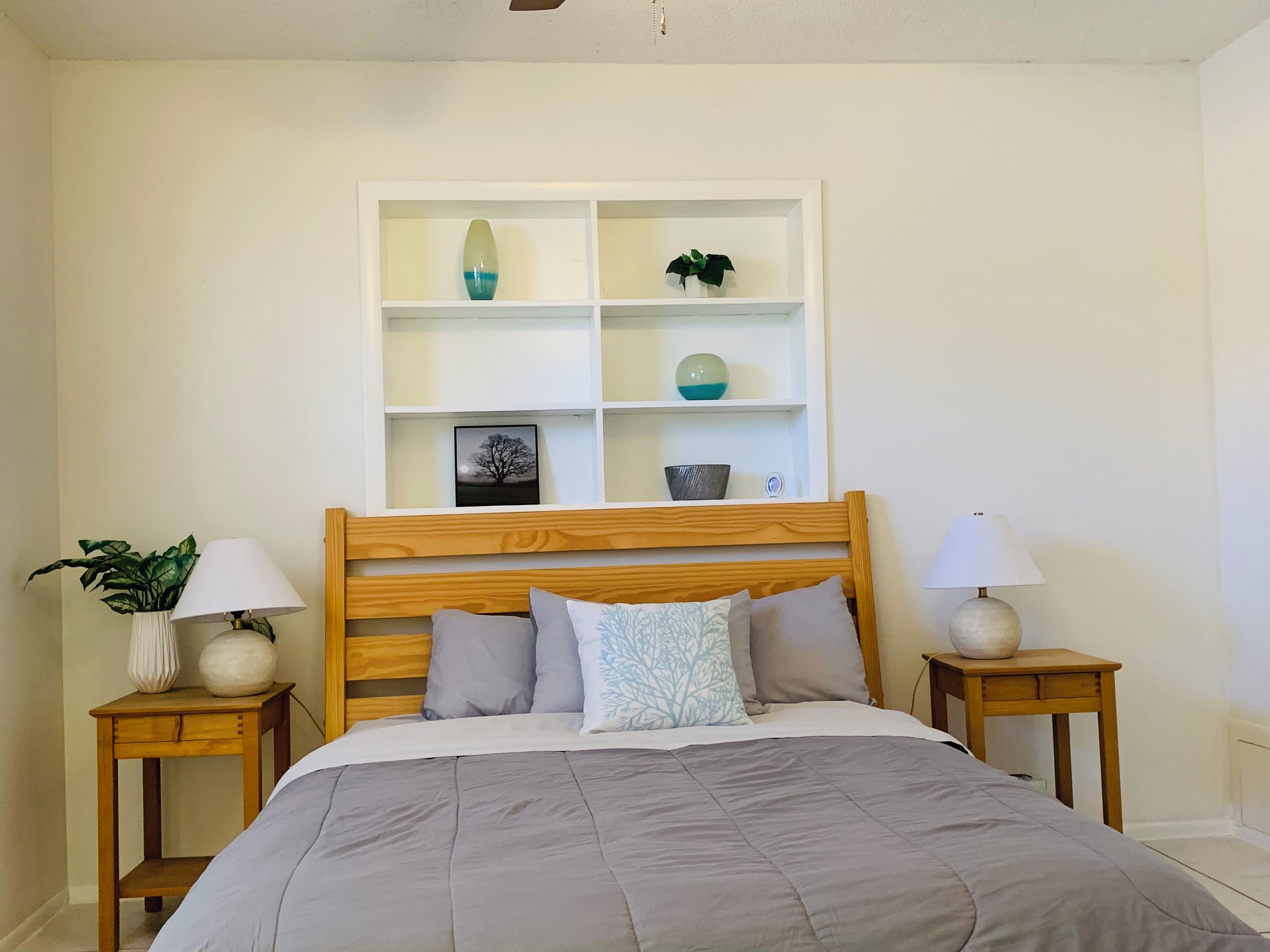 Un dormitorio con cabecera de madera, cama con ropa de cama gris, dos mesitas de noche con lámparas y un estante blanco sobre la cama con objetos decorativos.