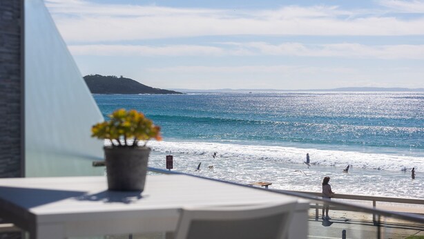 Ein Balkon mit einem Blumentopf, der Blick auf einen Strand mit Wellen und eine entfernte Insel gewährt.