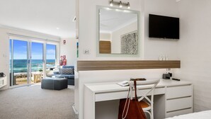 Ein Hotelzimmer mit einer großen Schiebeglastür, die auf einen Balkon mit Meerblick führt. Es gibt ein Bett, einen Schreibtisch mit Stuhl und einen an der Wand montierten Fernseher.