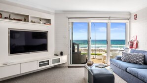 Ein modernes Wohnzimmer mit einem großen Flachbildfernseher, einer Couch und einem Glastisch. Eine Glasschiebetür führt zu einem Balkon mit Blick auf Strand und Meer.