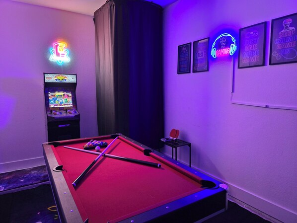 A pool table with a red felt surface, a pool cue, and several balls. There is a neon sign on the wall and a small table with a chair.