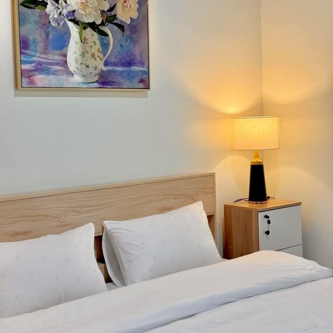 Habitación de hotel con cabecera de madera, cama con ropa blanca, mesita de noche con lámpara y un cuadro de flores en la pared.