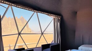 A room with a bed, two chairs, a small table, and a view of a desert landscape through a large window. - Siwar Luxury Camp  (Wadi Rum)