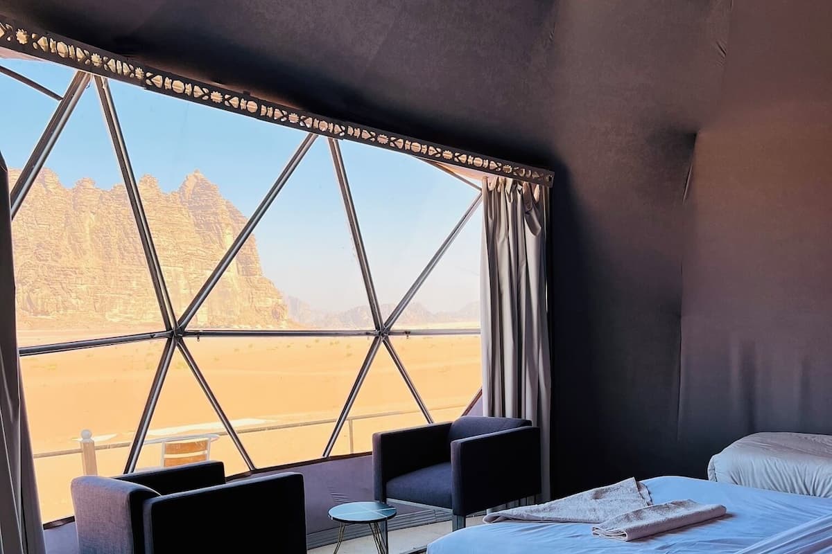 Une pièce comprenant un lit, deux chaises, une petite table et une vue sur un paysage désertique, visible à travers une grande fenêtre.