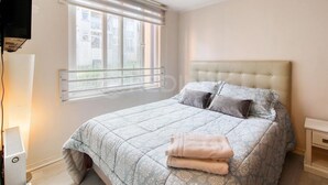 Quarto com cama, janela, mesa de cabeceira e televisão fixada na parede.