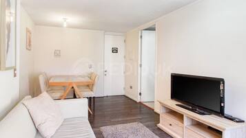 A modern living room with a sofa, a TV on a wooden stand, a dining table with chairs, and a door leading to another room.