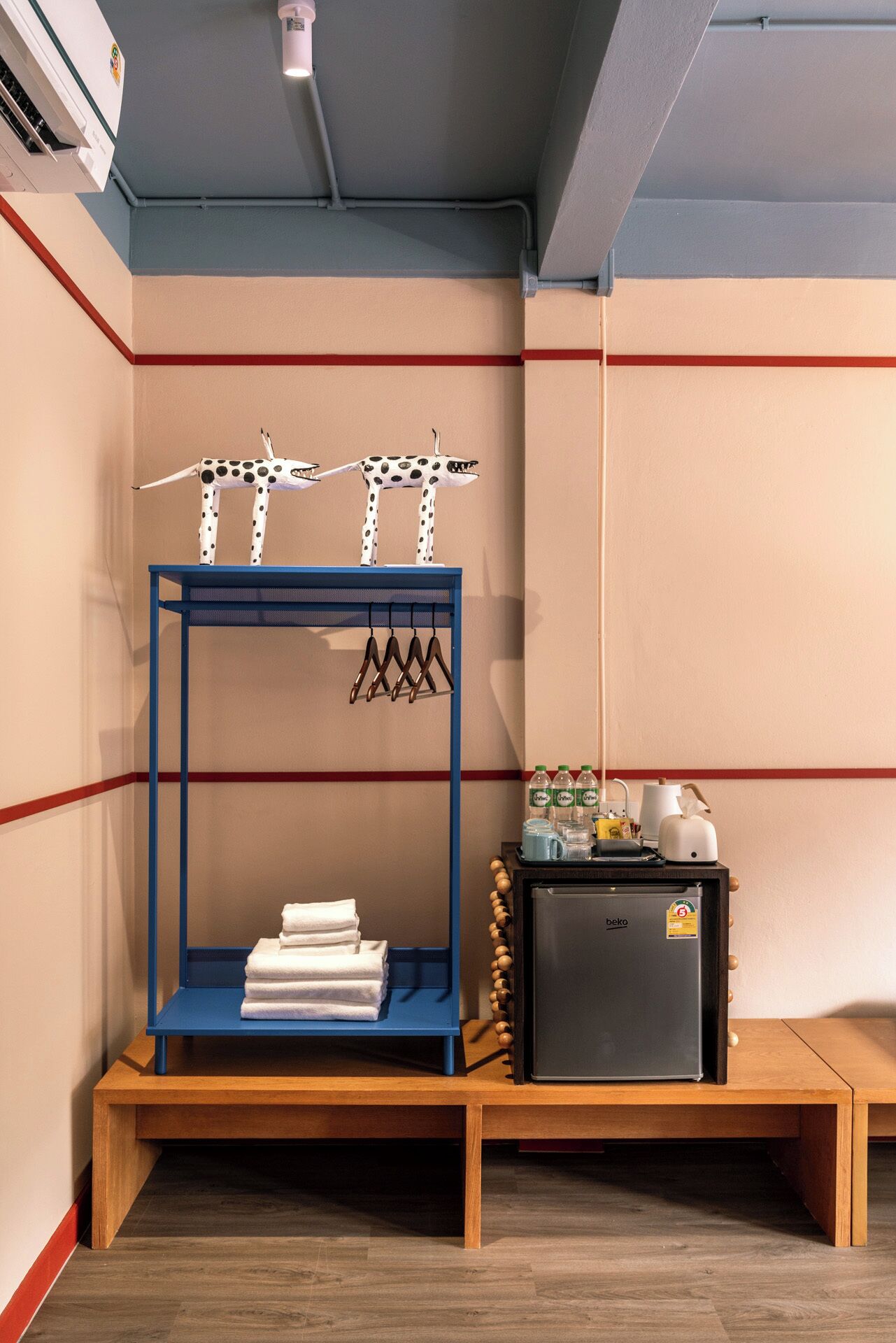 A compact room with a blue metal shelf unit holding towels and hangers, a small refrigerator with beverages, and a wall-mounted air conditioning unit.