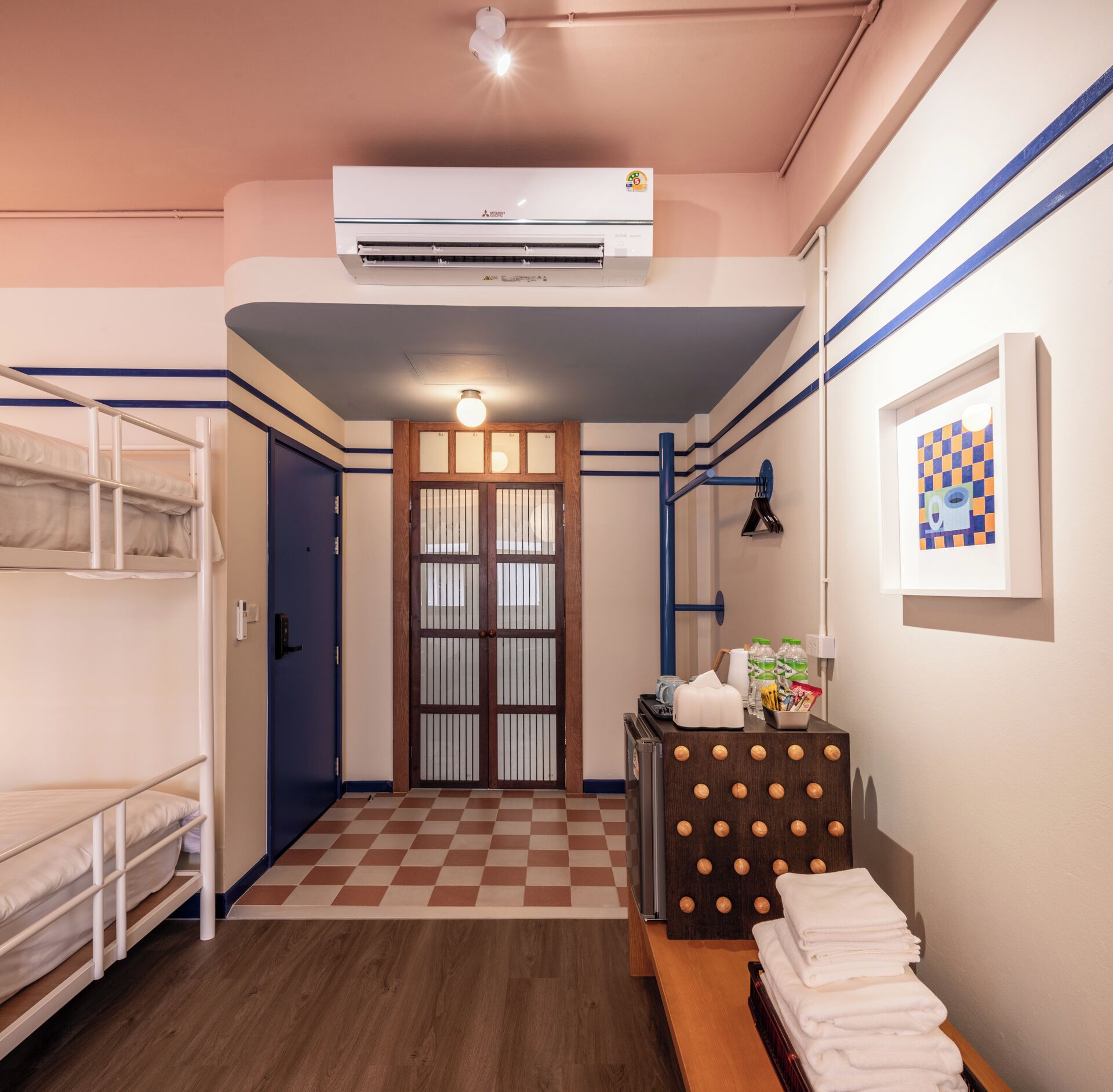 A hotel room with bunk beds, a checkered floor, an air conditioning unit, a door with a glass panel, and a wall-mounted shelf with towels.