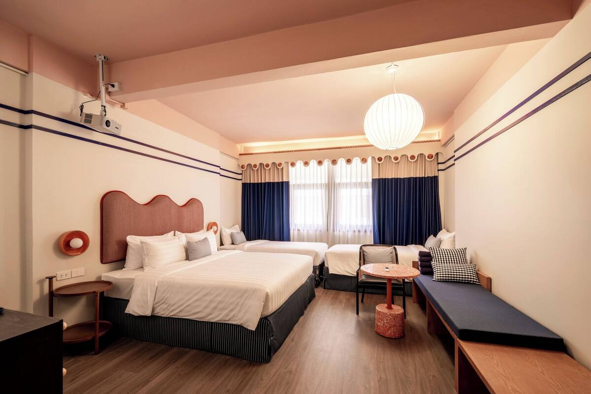 A hotel room with two beds, a small table, a bench, and a ceiling-mounted projector.