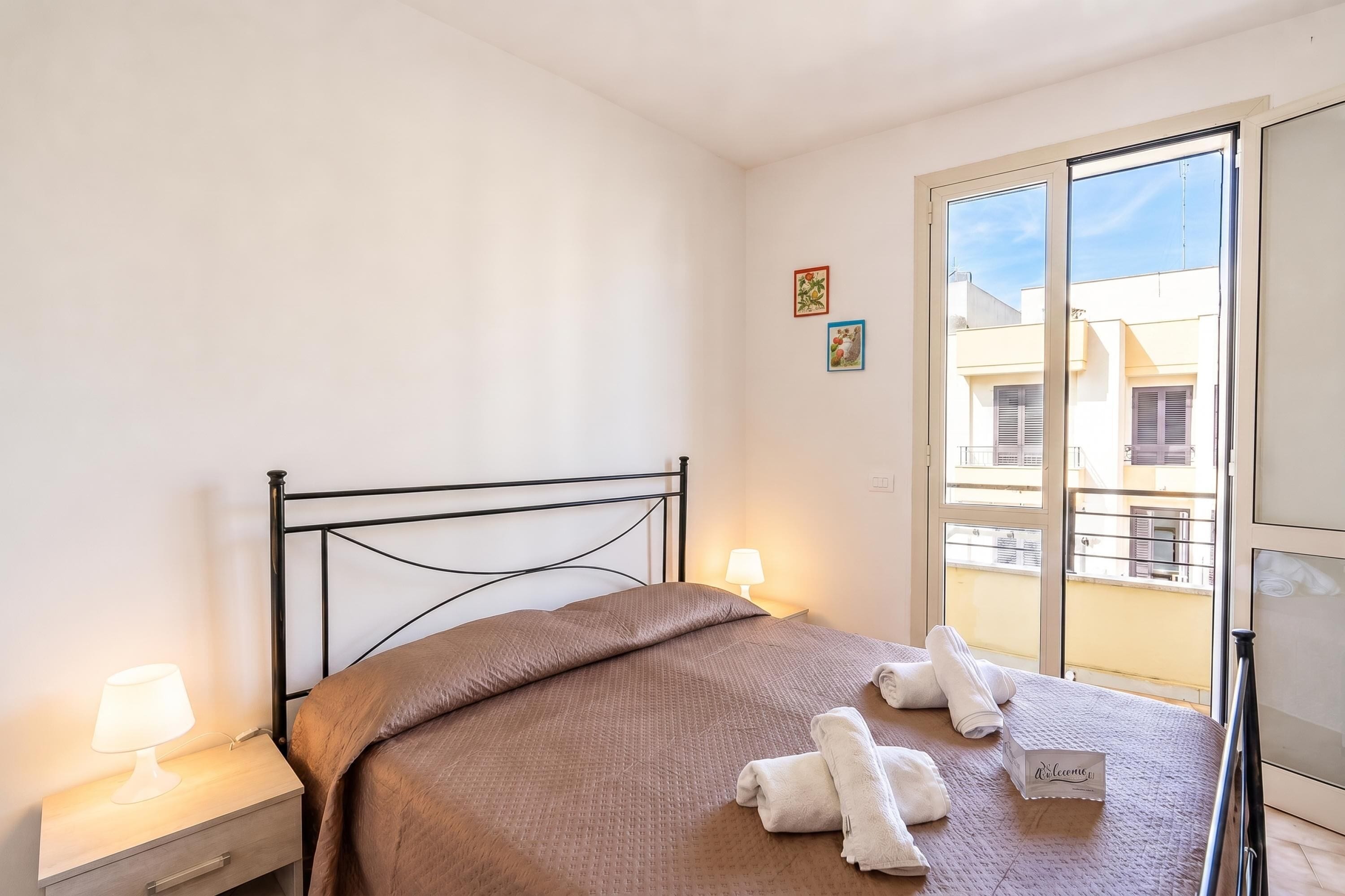 Une chambre d’hôtel avec un lit, une table de chevet, une lampe, un balcon donnant sur des immeubles et une porte.